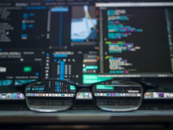 Zdjęcie przedstawiające okulary leżące na biurku, a w tle monitor z aplikacjami wyświetlającymi kod programu.