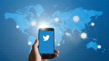 Zdjęcie ilustracyjne- ikona Twitter w telefonie, w tle  na niebiesko mapa świata z zaznaczonymi na biało punktami.