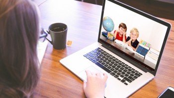 Zdjęcie ilustracyjne, które przestawia laptop położony na stole, na jego ekranie widoczne są dwie postaci dzieci, obok których stoją globus oraz podręczniki szkolne. Przed laptopem widoczna postać nauczyciela.