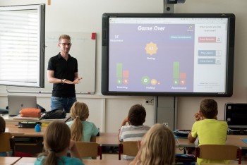 Zdjęcie przedstawia nauczyciela, który stoi obok tablicy interaktywnej i    
 mówi do uczniów.