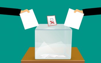 Obrazek ilustrujący urnę wyborczą, do której wrzucane są karty wyborcze.  